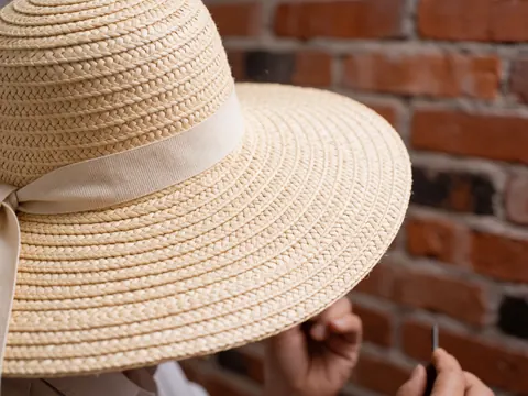 Gros plan d'un chapeau de paille orné d'un ruban beige, avec des mains travaillant sur un fond de mur en briques rouges.
