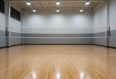 Vue d'un terrain de volley-ball intérieur vide, doté d'un sol en bois poli, d'un filet central et de murs gris avec zones de sécurité, idéal pour l'entraînement.