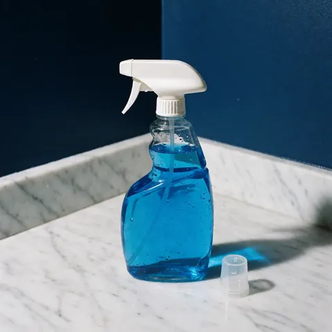 Bouteille de nettoyant bleu brillant avec vaporisateur sur surface en marbre blanc, couvercle à côté et mur bleu foncé en arrière-plan.
