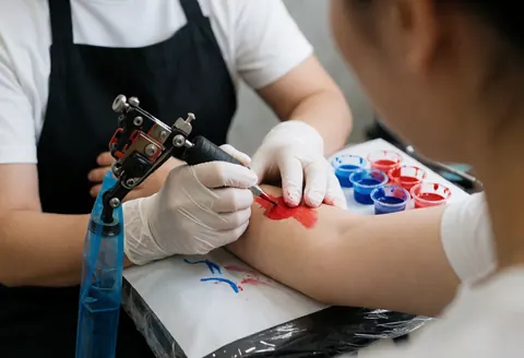 Un tatoueur portant des gants utilise une machine pour appliquer de l'encre rouge sur le bras d'un client, près d'une tablette avec des encres bleues et rouges dans un studio propre.