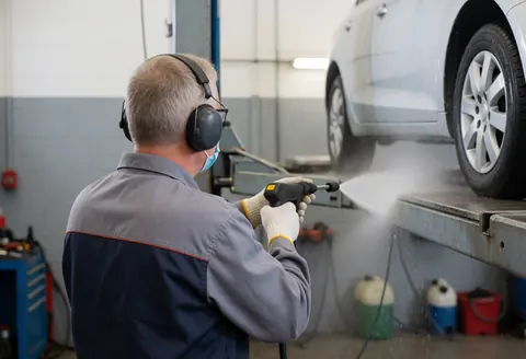 Un ouvrier en atelier automobile utilise un équipement de protection individuelle pour nettoyer un véhicule avec une lance à eau sous pression, entouré d'outils.