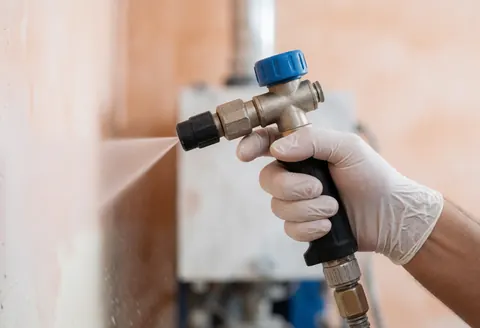 Une main portant un gant blanc guide un tuyau qui projette un jet d'eau sur un mur terracotta, avec du matériel industriel en arrière-plan flou.