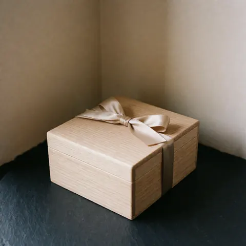 Boîte en bois ornée d'un ruban beige posée sur une surface sombre dans le coin d'un mur beige, éclairage doux et naturel.