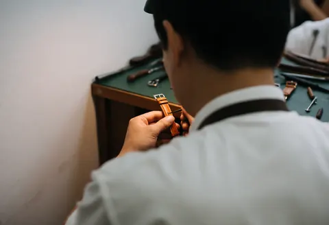 Un horloger en chemise blanche travaille dans son atelier en appliquant un soin sur une courroie de montre en cuir marron posée sur une table verte.
