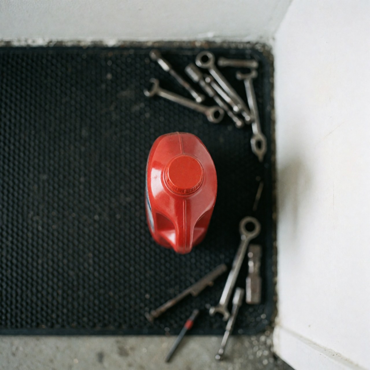 Vue de dessus d'une bouteille rouge contenant du liquide, posée sur un tapis noir avec des clés à rotule dispersées autour dans un atelier mécanique.