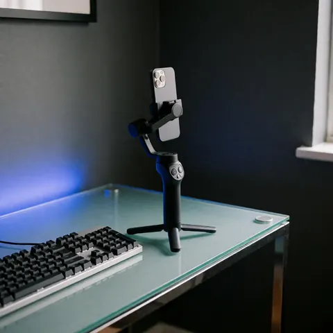 Un gimbal noir posé à côté d'un smartphone sur un bureau en verre transparent, près d'un clavier, éclairé par une lumière bleue dans un environnement minimaliste aux murs gris.