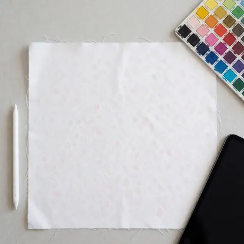 Tissu blanc imprimé numériquement, palette de couleurs et stylet posés sur un fond gris clair, éclairage uniforme, scène de design graphique.