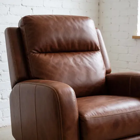 Un fauteuil en cuir marron restauré avec des coutures visibles et une texture riche, posé dans un atelier devant un mur de briques blanches.
