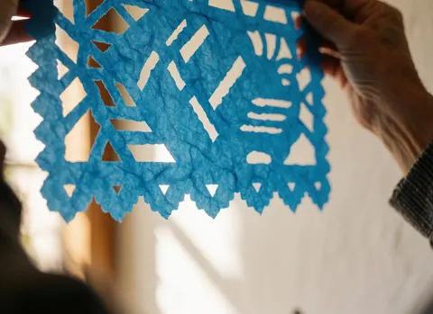 Près d'un drapeau en papier picado bleu, mains tenant le détail, lumière du soleil filtrant à travers les coupes, texture visible du papier.