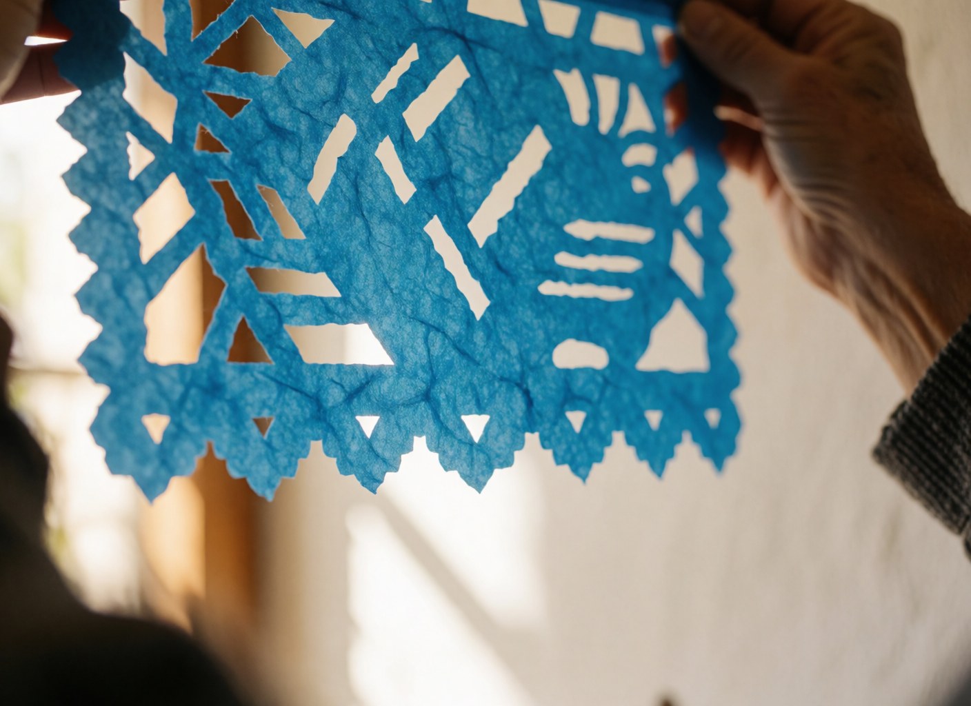 Près d'un drapeau en papier picado bleu, mains tenant le détail, lumière du soleil filtrant à travers les coupes, texture visible du papier.