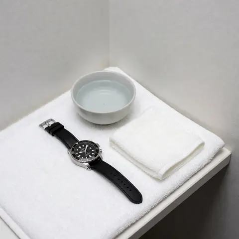 Montre-bracelet à bracelet noir sec posée sur une serviette blanche, accompagnée d'une tasse d'eau et d'un chiffon, dans un coin aux murs gris clair, avec un éclairage lumineux et propre.