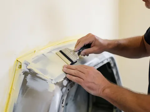 Une main utilise une spatule pour appliquer uniformément du mastic sur une surface métallique de voiture, préparée avec du ruban jaune sur un fond blanc.