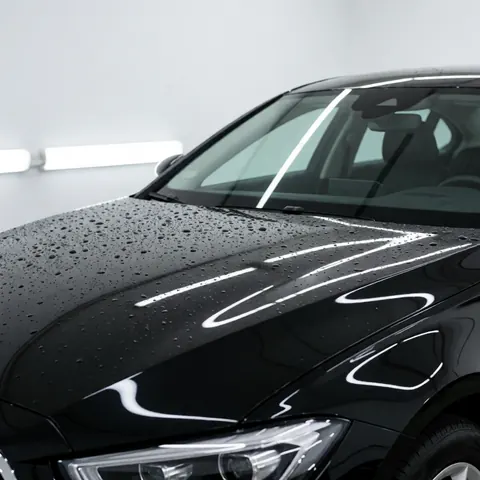 Capot de voiture noir avec finition céramique, des gouttes d'eau reflètent la lumière LED sur un fond blanc dans un atelier de detailing automobile.