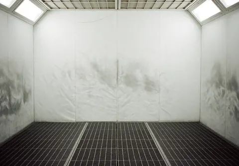 Intérieur d'une cabine de peinture automobile professionnelle vide, dotée d'un sol en grille métallique et de murs blancs tachés de peinture, éclairée par des luminaires au plafond.