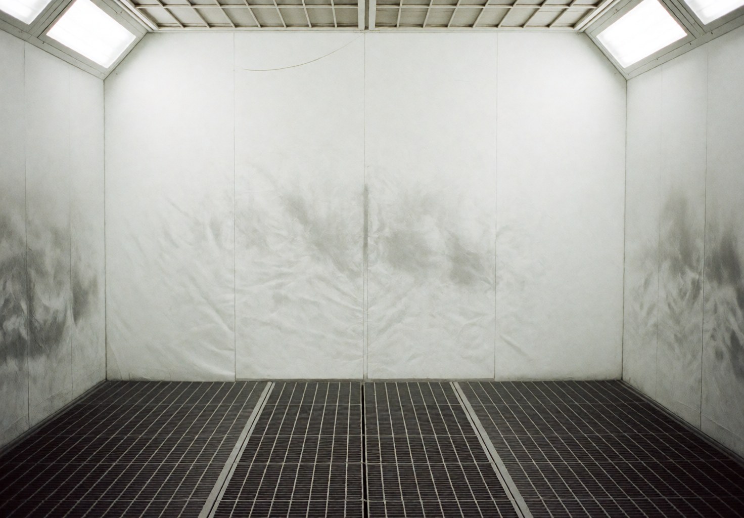 Intérieur d'une cabine de peinture automobile professionnelle vide, dotée d'un sol en grille métallique et de murs blancs tachés de peinture, éclairée par des luminaires au plafond.