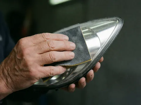 Une main adulte utilise du papier abrasif humide pour polir la surface courbe d'un phare de voiture sur fond sombre et flou, mettant en valeur la texture et le brillant.