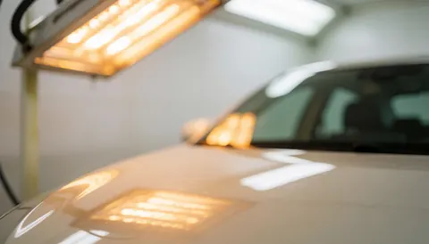 Capot de véhicule en cours de séchage sous éclairage infrarouge dans un atelier de carrosserie, avec un arrière-plan flou aux tons chauds et une ambiance professionnelle.