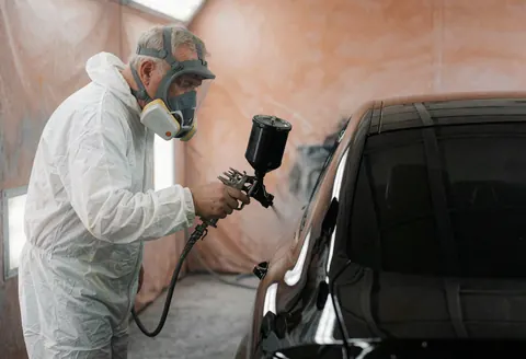 Un technicien en tenue blanche et masque peint le capot d'une voiture noire brillante dans une cabine avec des murs terracotta.