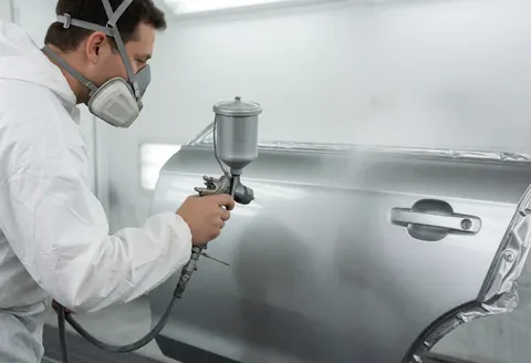 Un peintre en combinaison blanche et masque applique de la peinture métallique sur une portière dans un atelier. Fond blanc, lumière uniforme.