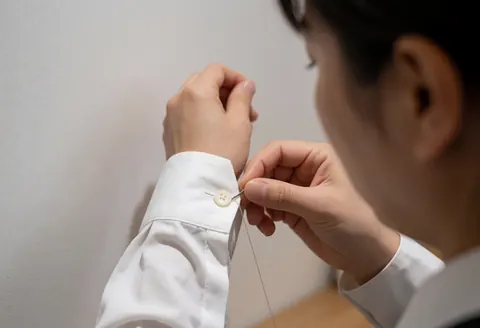Gros plan d'une main adulte en train de coudre un bouton sur la manche d'une chemise blanche, sur fond blanc avec éclairage naturel.