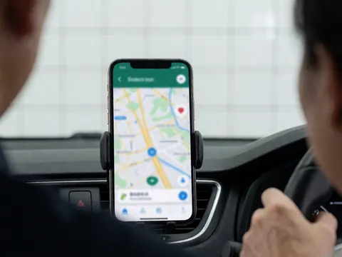 Un smartphone monté sur le tableau de bord affiche une carte de navigation. Les mains du conducteur sont posées sur le volant, devant un fond de murs carrelés blancs.