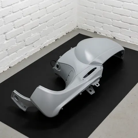 Panneau de carrosserie de scooter peint en gris mat posé sur une surface noire mate devant un mur de briques blanches avec un éclairage professionnel.