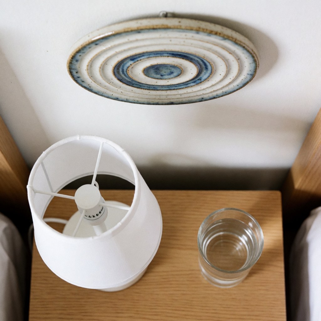 Table de chevet en bois avec lampe blanche, verre d'eau et art céramique au mur. Scène épurée et minimaliste pour chambre d'hébergement.