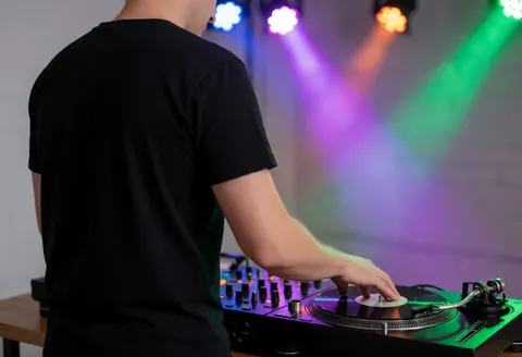 Un DJ de fêtes mixant de la musique sur une table de mixage avec des lumières de scène en arrière-plan, des murs gris et une ambiance de fête.