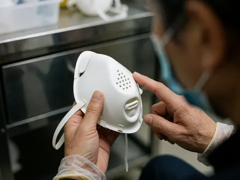 Un professionnel portant des gants examine de près un masque médical blanc équipé de valves respiratoires sur une surface métallique.