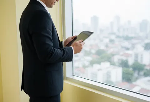 Un homme d'affaires en costume sombre tient une tablette tout en regardant par une fenêtre offrant une vue sur la ville, avec un mur jaune pâle en arrière-plan.