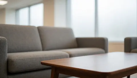 Fond professionnel de salle de vente avec canapé gris texturé et table en bois, lumière diffuse par les fenêtres, idéal pour textes commerciaux.