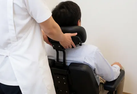 Un praticien en tablier blanc applique un massage shiatsu sur le cou d'un client assis dans une chaise ergonomique noire, dans un environnement professionnel et apaisant.