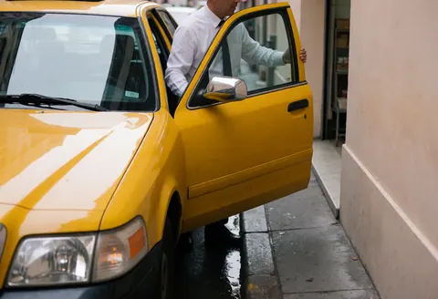 Chauffeur de taxi jaune debout près de sa voiture avec la portière ouverte dans une rue urbaine humide sous un ciel nuageux.