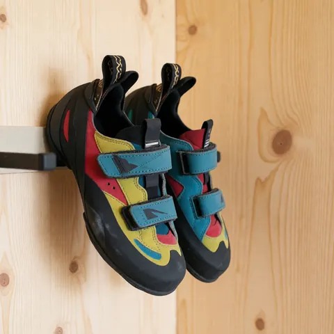 Des chaussures de grimpe aux couleurs vives avec des lacets velcro, accrochées à un support mural en bois clair éclairé par la lumière naturelle.