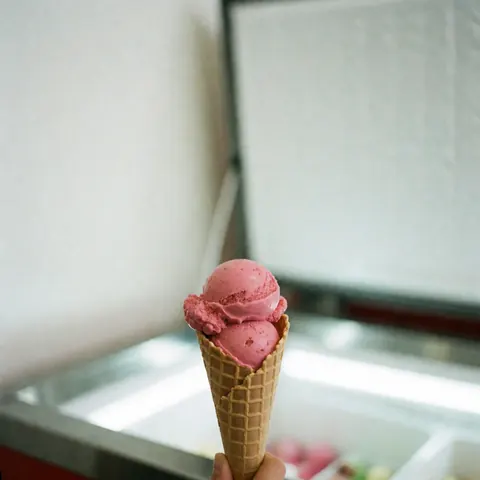 Une glace à la fraise rose sur un cône en gaufre est tenue devant une vitrine de glacier avec un fond blanc et une lumière vive.
