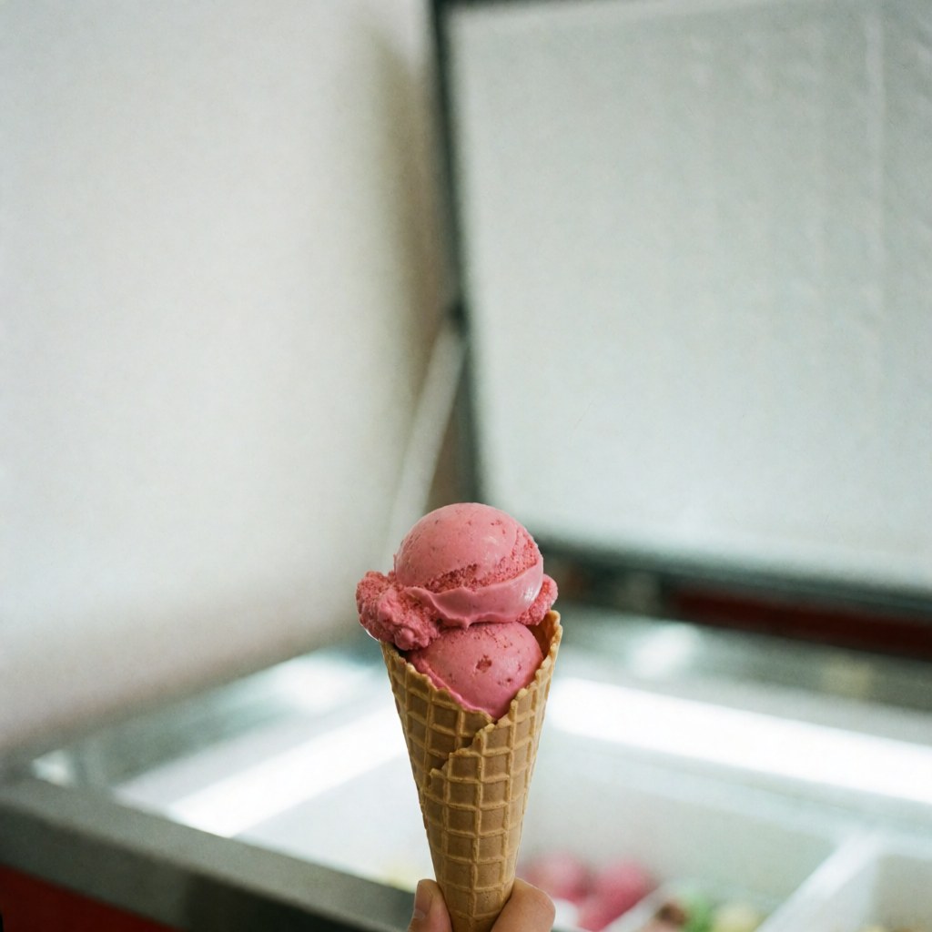 Une glace à la fraise rose sur un cône en gaufre est tenue devant une vitrine de glacier avec un fond blanc et une lumière vive.