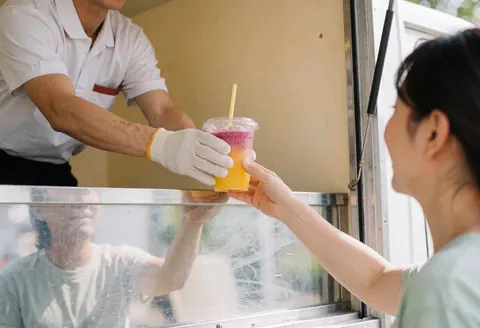 Un employé en uniforme et gants remet une boisson colorée à un client dans un camion de vente ambulante.