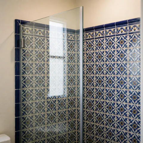 Paroi de douche transparente sans cadre installée sur un mur recouvert de carreaux Talavera traditionnels bleus et blancs à motifs décoratifs, sous un éclairage doux.