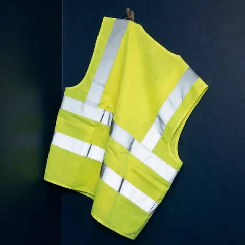 Un gilet de sécurité jaune avec des bandes réfléchissantes est accroché sur un mur bleu marine, éclairé par une lumière chaude qui met en valeur ses détails et sa texture.