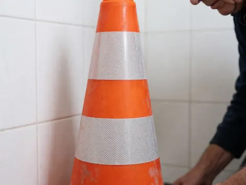 Main d'un travailleur ajustant un cône de sécurité orange à bandes blanches réfléchissantes devant un mur de carreaux blancs.