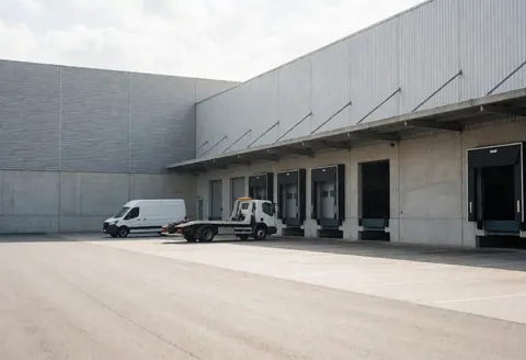 Un remorqueur blanc et une camionnette sont garés devant des quais de chargement dans une vaste zone industrielle vide, sous un éclairage naturel sur un sol en béton.