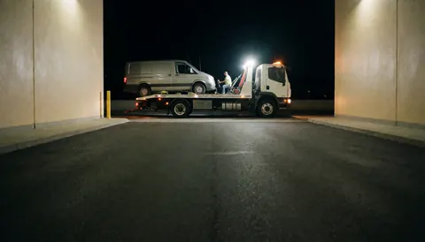 Un remorqueur blanc avec une van sur sa plateforme, éclairé par des projecteurs puissants dans un espace pavé bordé de murs beige.