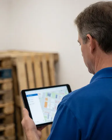 Un homme en chemise bleue consulte une carte sur une tablette. En arrière-plan, des palettes en bois sont empilées dans l'entrepôt, illustrant une vérification logistique.