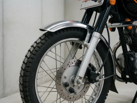 Gros plan sur l'avant d'une moto de transport urbain montrant la roue, la suspension, le pneu tout-terrain et le garde-boue chromé contre un mur en béton.