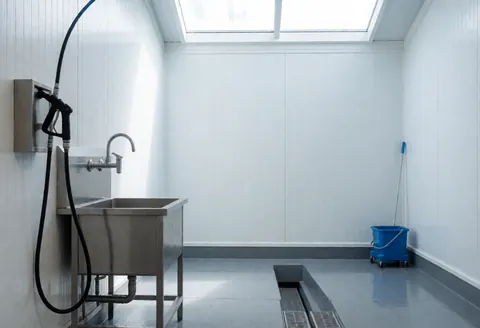 Espace de nettoyage pour bicyclettes équipé d'un robinet, d'un tuyau haute pression et d'une canalisation au sol gris. Murs blancs et lumière naturelle provenant du plafond.