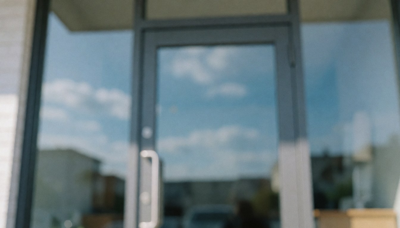 Porte d'entrée vitrée à cadre gris reflétant un ciel bleu nuageux, offrant un fond neutre et flou idéal pour l'intégration de textes commerciaux.