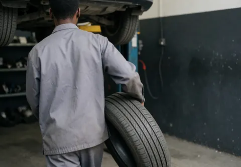 Mécanicien de dos transportant un pneu neuf vers un véhicule sur élévateur dans un atelier avec étagères de pièces en arrière-plan