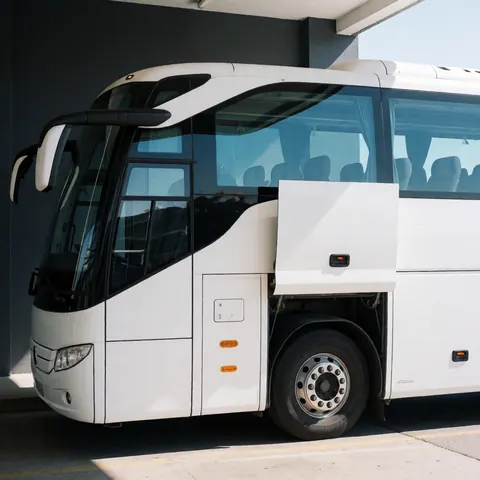 Un bus touristique blanc est stationné dans un terminal aux murs gris. Les vitres teintées et les roues sont visibles sous la lumière du soleil.
