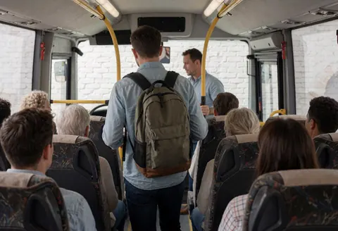 Un voyageur équipé d'un sac à dos monte dans un autocar. Un guide l'assiste. Des sièges et des murs en briques blanches sont visibles dans l'habitacle.