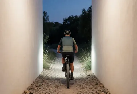 Un cycliste en tenue technique et casque pédale sur un sentier caillouteux bordé de murs blancs au crépuscule, son phare éclaire la route.